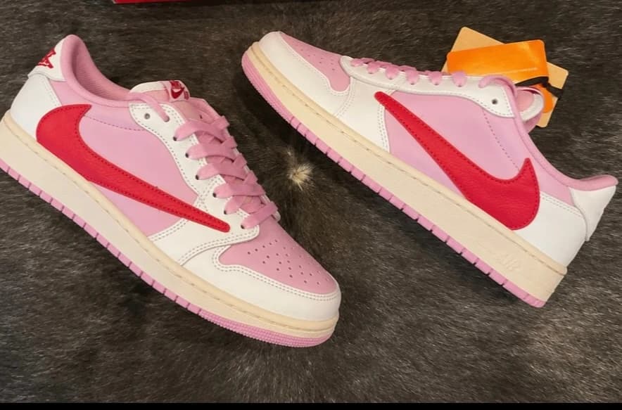 Um par de Air Jordan Low 1 Travis Scott Tropical Pink, rosa com o swoosh vermelho invertido, no tapete da loja